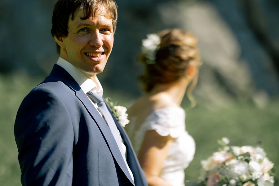 Nahaufnahme des Bräutigams der in die Kamera lächelt, im Hintergrund kann man die Braut erkennen.